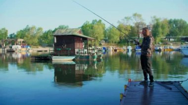 Uzun saçlı bir adam salla nehir kenarında balık tutuyor. Etrafında kayıklar ve yüzen evler ve arka planda ağaçlar var. Balıkçı oltası kullanıyor, makaralı, yeşil ipli ve yemli..