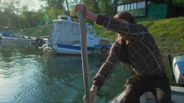 Beyaz orta yaşlı adam eski beyaz teknesinde kürek çekiyor. Gün doğumunda nehir marinasında. Bu balıkçı eski tahta küreği kullanıyor. Etrafında kayıklar ve yüzen evler ve arka planda ağaçlar var..