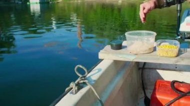 Balık yemi ve balık yemi için yemek yapan eller. Adam mısır ekleyip plastik kaseye karıştırıyor. Beyaz bir kayıkla nehirde yüzüyor ve tahta bir tahtanın üzerinde oturuyor..
