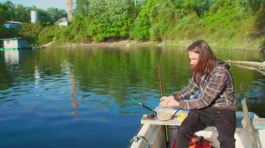 Balıkçı, yemi göle atıyor. Etrafında ağaçlar olan beyaz bir teknede. Adamın uzun kahverengi saçları var. İnşaat alanı arka planda. Mısır ve solucanlar su yüzeyine sıçrıyor damlalara neden oluyor..