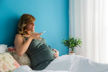 Hamile bir kadın, akıllı telefonuyla karnının fotoğraflarını çekerken yatağında yarı oturuyormuş..