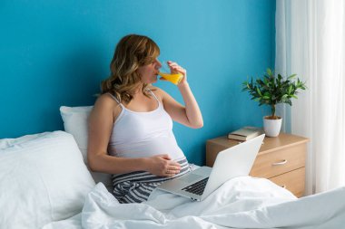 Dizüstü bilgisayarı üzerinde çalışırken yatağında oturan gülümseyen ve mutlu, hamile, beyaz bir kadın. Bir eli karnında ve meyve suyu içiyor.