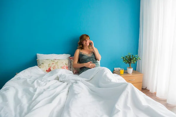 A pregnant woman in a semi-sitting position in her bed while talking over her smart phone. She is looking happy with one hand on her stomach.