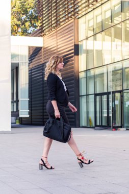 Modern bir binanın önünde elinde laptop çantasıyla yürüyen hamile bir iş kadını..