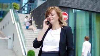 Modern binanın önünde bavulla yürüyen ve cep telefonundan bir şeyler okuyan gülümseyen hamile bir iş kadını. Arkaplan, perdeli büyük cam pencerelerden oluşur.