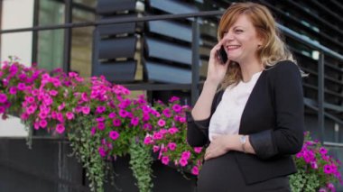 Modern bir binanın önünde çiçeklerin yanında cep telefonuyla konuşan gülümseyen hamile bir iş kadını..