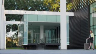 Modern bir binanın önünde elinde bir parça seyahat valiziyle yürüyen hamile, beyaz bir kadın kol saatine bakıyor..