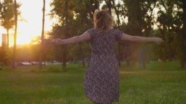 Günbatımında arka planda ağaçlarla şehir parkında duran gülümseyen beyaz hamile bir kadın. Elbisesini tek eliyle tutuyor..