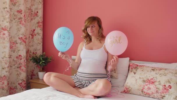 一个笑着 快乐地怀孕的高加索女人 拿着两个气球坐在床上 她拿气球开玩笑 上面写着 是个男孩 是个女孩 — 图库视频影像