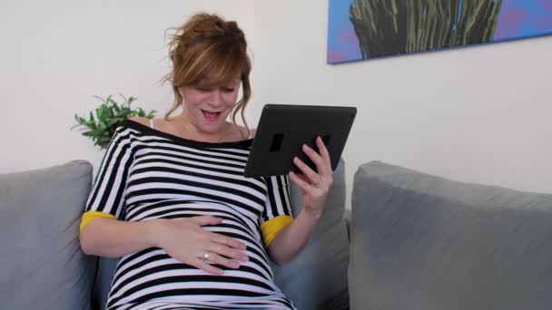 ソファの上の半座の位置に笑顔の妊婦は 彼女のタブレット上で何かを読み取ります お腹の赤ちゃんのキックに笑顔で反応し 胃を片手に — ストック動画