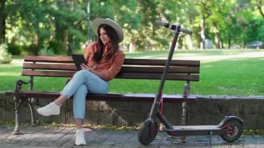 Şapkalı genç bir kadın parkta bir bankta oturuyor ve elindeki tabletin üzerinde kayıyor. Arkaplanda ağaçlar ve yeşil renkler baskın iken, onun yanında bir elektrikli scooter park edilir.