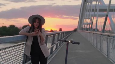 Genç, gülümseyen bir kadın köprü çitlerine yaslanıyor ve telefonuna yazı yazıyor. Onun yanına elektrikli bir scooter park edilmiş. Nehir, günbatımı ve arka planda modern bir köprünün parçaları yer alıyor..