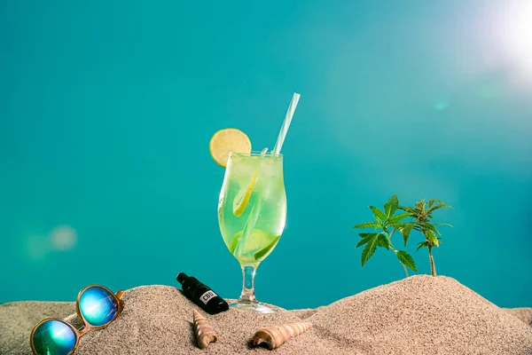 Farklı bardaklarda limonata, CBD damlası yağı ve kumda bir dal marihuana. Güneş gözlüğü ve deniz kabukları yan taraftan görülebilir. Arka plan yarı kumda, yarı mavi renkte..