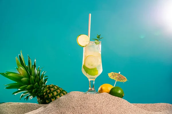 Refreshing jar/glass of lemonade and pineapple in the sand. Also, pineapple, lemon and lime can be seen around. The background is half in the sand while the other half is in the form of a blue wall.