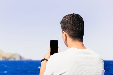 Maghreb kökenli tanınmayan koyu renk saçlı genç adam, arkasından akıllı telefonuyla plajın dikey fotoğrafını çekiyor. Güneşli ve aydınlık yaz günü, açık ve mavi renkler.