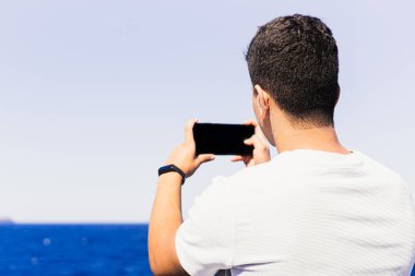 Maghreb kökenli tanınmayan koyu renk saçlı genç adam, arkasından akıllı telefonuyla kumsalın yatay fotoğrafını çekiyor. Güneşli ve aydınlık yaz günü, açık ve mavi renkler.