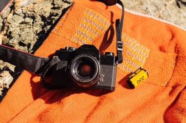 Old unrecognizable black analog photo camera on an orange background with a 35mm reel film next to it. On top of some sea rocks on a sunny day in Spain. clipart
