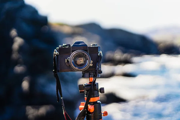 Black unbranded reflex (analog) camera planted on a tripod facing the camera. With a seascape in the background on a sunny summer day.