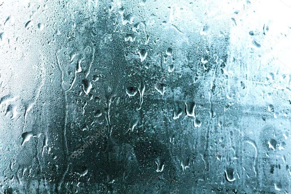Foggy glass of a car window with drops falling on a rainy day, blue background.