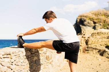 Genç sporcu, deniz kenarına bakarken bacaklarını dışarı uzatarak ufka bakıyor. Fotoğraf, çok güneşli bir günde Akdeniz manzaralı bir arkadan çekildi..