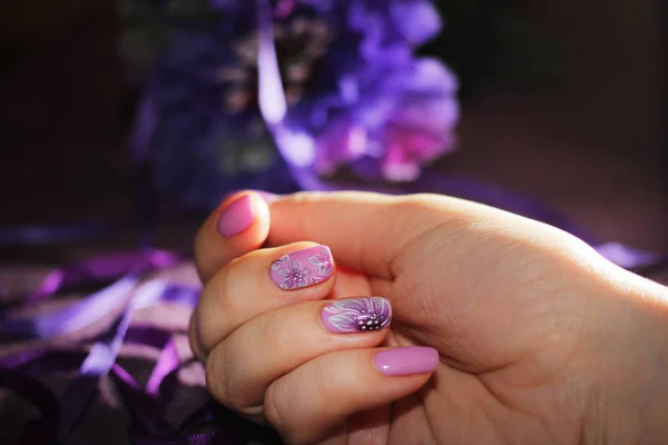 Purple design of nails with drawings of flowers. In the hand a purple ribbon and in the background a flower in the tone of a manicure.