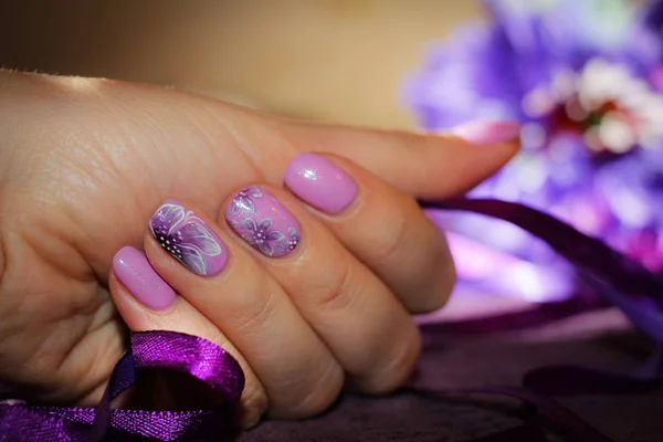 Purple design of nails with drawings of flowers. In the hand a purple ribbon and in the background a flower in the tone of a manicure.