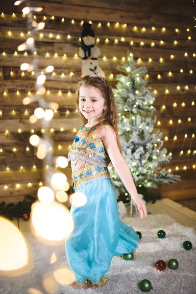 beautiful Caucasian little girl in blue costume in Christmas house dancing, garland with lights on wooden wall