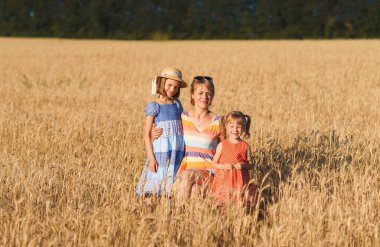 Anne, güneşli bir günde buğday tarlasında iki kıza sarılıyor.