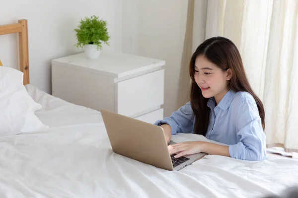 CUte asian girl freelancer lying on the bed working on a laptop, having breakfast in bed. Work from home, stay home concept, home office, working home, self-isolation.