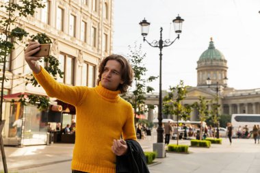 Genç dostum bekleyen biri ve selfie yapar, o sarı kazak, siyah yağmurluk ya da ceket, kot pantolon, sokak ve arka plan üzerinde Kazanskiy Katedrali giyinmiş güneşli bir gün