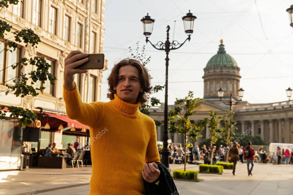 The young man waiting someone and does selfie, he dressed in a yellow sweater, a black raincoat or jacket, jeans, street and Kazanskiy cathedral on background, sunny day