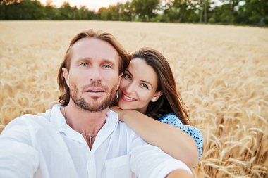 Güzel çift bir çavdar alanında kucaklayan, bir kamera, güzel gözler, mutlu gülümsüyor, uzun saç, kolay bir sakal, arka plan, mavi elbise ve beyaz gömlek buğday kulaklara görünüyor