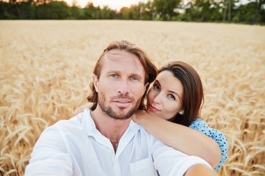 Güzel çift bir çavdar alanında kucaklayan, bir kamera, güzel gözler, mutlu gülümsüyor, uzun saç, kolay bir sakal, arka plan, mavi elbise ve beyaz gömlek buğday kulaklara görünüyor