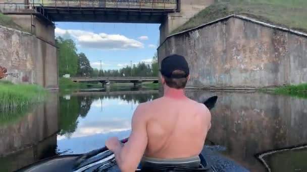 Ryssland, Gatchina, 29 maj 2020: De unga männen i en mössa flyter på en kajak under bron, det vackra landskapet, den lugna floden, ett soligt väder, aktivt ror med en åra — Stockvideo