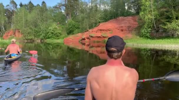 Rusia, Gatchina, 29 de mayo de 2020: Los dos jóvenes en una gorra flotan en un kayak en el río del bosque, el hermoso paisaje, el río tranquilo, un clima soleado, reman activamente con un remo, costa Ocherous — Vídeos de Stock