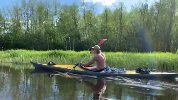 Rosja, Gatchina, 29 maja 2020 r.: Młodzi mężczyźni w czapce pływają kajakiem po leśnej rzece, pięknym krajobrazie, cichą rzeką, słoneczną pogodą, aktywnie hałasują wiosłem, piękną refleksją — Wideo stockowe