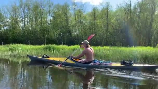 Rússia, Gatchina, 29 de maio de 2020: Os jovens em um gorro flutuam em um caiaque no rio de floresta, a bela paisagem, o rio tranquilo, um tempo ensolarado, ativamente rema com um remo, bela reflexão — Vídeo de Stock