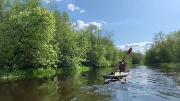 Rússia, Gatchina, 29 de maio de 2020: Os jovens em um gorro flutuam em um caiaque no rio de floresta, a bela paisagem, o rio tranquilo, um tempo ensolarado, ativamente rema com um remo, bela reflexão — Vídeo de Stock