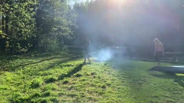 Rússia, Gatchina, 29 de maio de 2020: Os homens arrasta um log para lareira, lenha para um incêndio, a costa do rio, caras fizeram uma parada e caiaques secos, tempo ensolarado, cor verde, fumaça de fogo, gramado verde — Vídeo de Stock