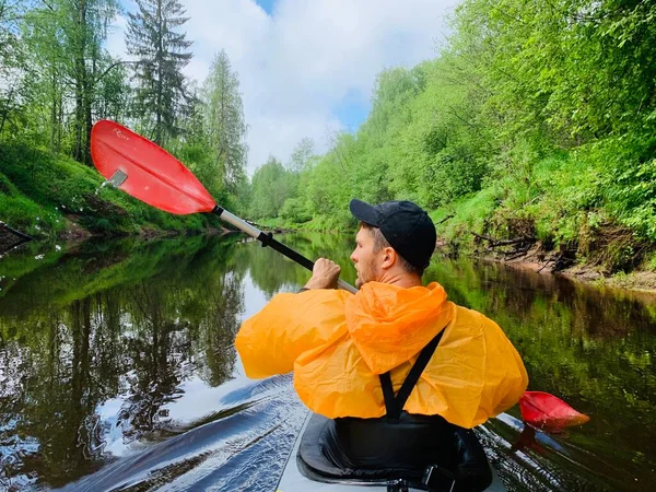 Russland, Kirishi, 25. Mai 2019: Die Männer in Mütze und orangefarbenem Regenmantel schwimmen auf einem Kajak auf dem ruhigen Fluss des Waldes, die schöne Landschaft, das wechselhafte Wetter, rudern aktiv mit dem Ruder. — Stockfoto
