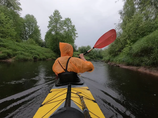 The men in a raincoat of orange color floats on a kayak on the forest quiet river, the beautiful landscape, a rainy weather, actively rows with an oar, beautiful reflection — Stock Photo, Image