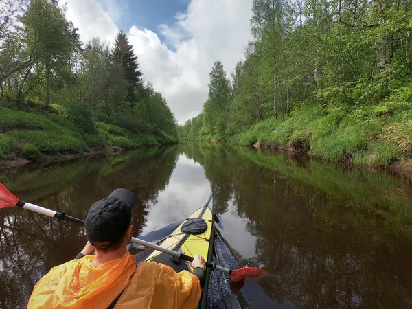 Die Männer in Mütze und orangefarbenem Regenmantel schweben auf einem Kajak auf dem ruhigen Fluss des Waldes, die schöne Landschaft, das wechselhafte Wetter, aktiv rudert mit einem Ruder, schönes Spiegelbild — Stockfoto