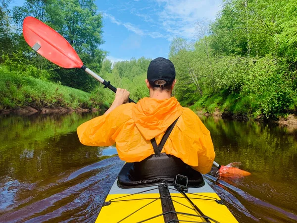 The men in a cap and raincoat of orange color floats on a kayak on the forest quiet river, the beautiful landscape, a changeable weather, actively rows with an oar, beautiful reflection — Stock Photo, Image