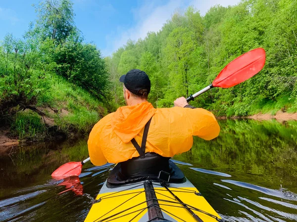 The men in a cap and raincoat of orange color floats on a kayak on the forest quiet river, the beautiful landscape, a changeable weather, actively rows with an oar, beautiful reflection — Stock Photo, Image