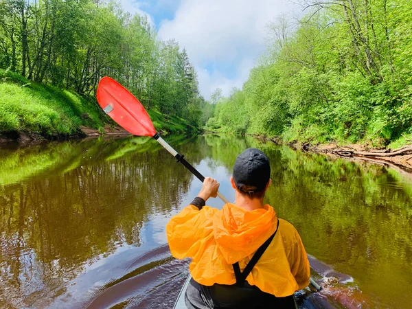 Die Männer in Mütze und orangefarbenem Regenmantel schweben auf einem Kajak auf dem ruhigen Fluss des Waldes, die schöne Landschaft, das wechselhafte Wetter, aktiv rudert mit einem Ruder, schönes Spiegelbild — Stockfoto