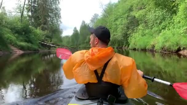Ryssland, Kirishi, 25 maj 2019: Männen i mössa och regnrock i orange färg flyter på en kajak på den lugna skogsfloden, det vackra landskapet, ett skiftande väder, aktivt radar med en åra — Stockvideo