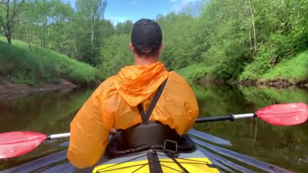 Rusland, Kirishi, 25 May 2019: Mændene i en kasket og regnfrakke af orange farve flyder på en kajak på den rolige skovflod, det smukke landskab, et foranderligt vejr, rækker aktivt med en år. instrukt – Stock-video