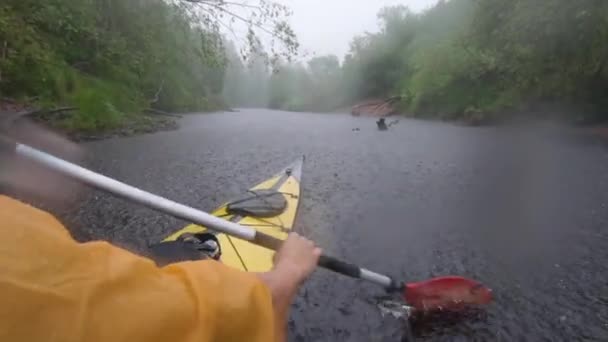 Rusia, Kirishi, 25 de mayo de 2019: Los hombres en un impermeable de color naranja flotan en un kayak en el bosque río tranquilo, el hermoso paisaje, un clima lluvioso, reman activamente con un remo — Vídeos de Stock