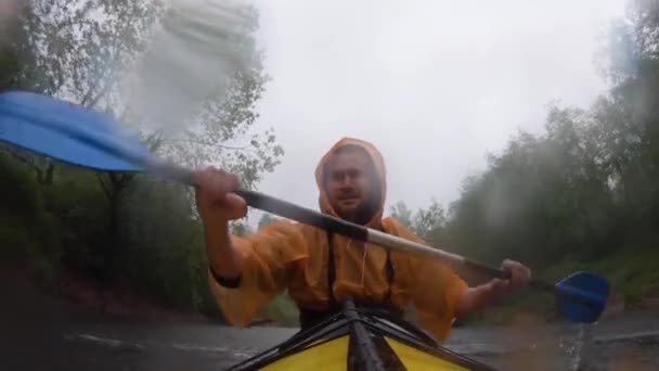 Rusia, Kirishi, 25 de mayo de 2019: Los hombres en un impermeable de color naranja flotan en un kayak en el río tranquilo bosque, la lluvia cae sobre una lente, un clima lluvioso, reman activamente con un remo — Vídeo de stock