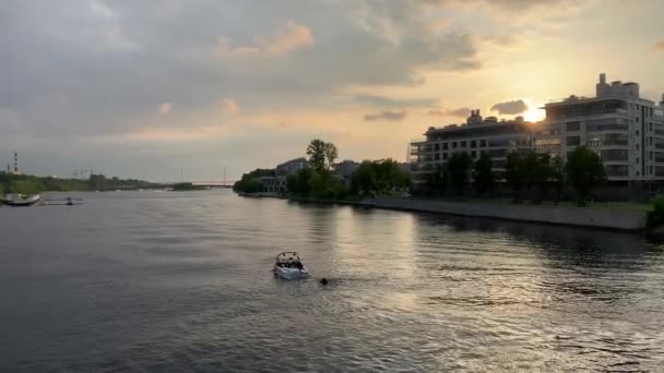 Ryssland, St Petersburg, 10 juni 2020: Båten med en väckarklocka på en våg går nerför floden vid solnedgången, ett bostadsområde på en bakgrund, solfläckar av ljus i fönster i byggnader — Stockvideo